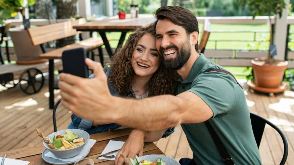 A man and woman smile while taking a selfie at an outdoor restaurant table.