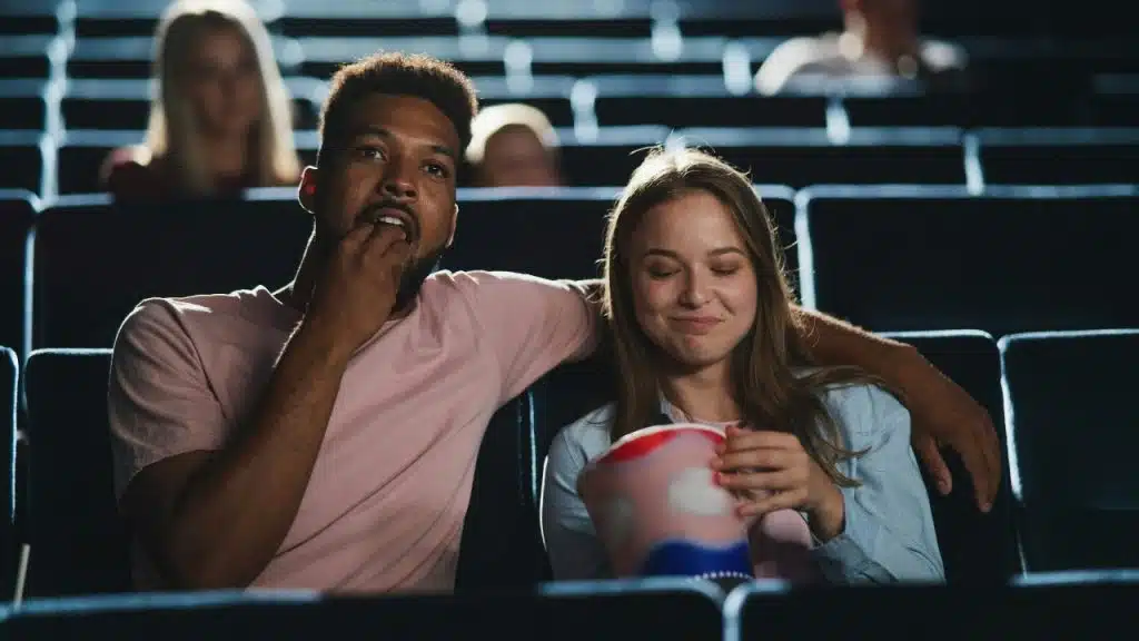 A man and woman sit in a dark theater eating popcorn while watching a movie.
