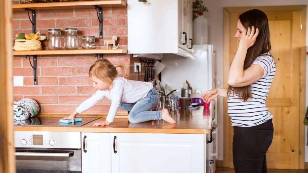A small child cleans a kitchen counter with a sponge while a woman stands nearby.