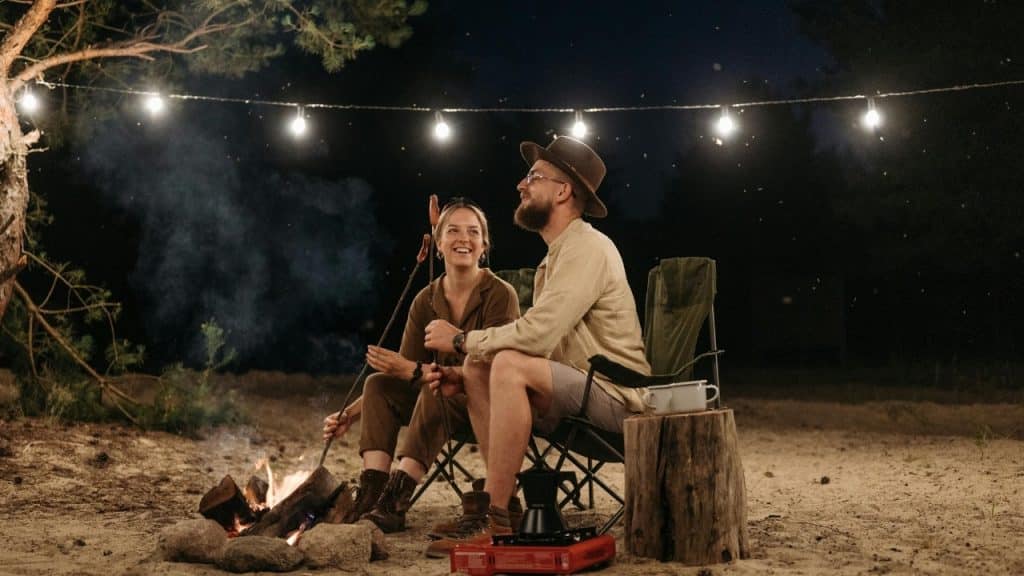 Two people sit in chairs by a campfire at night under a string of lights.