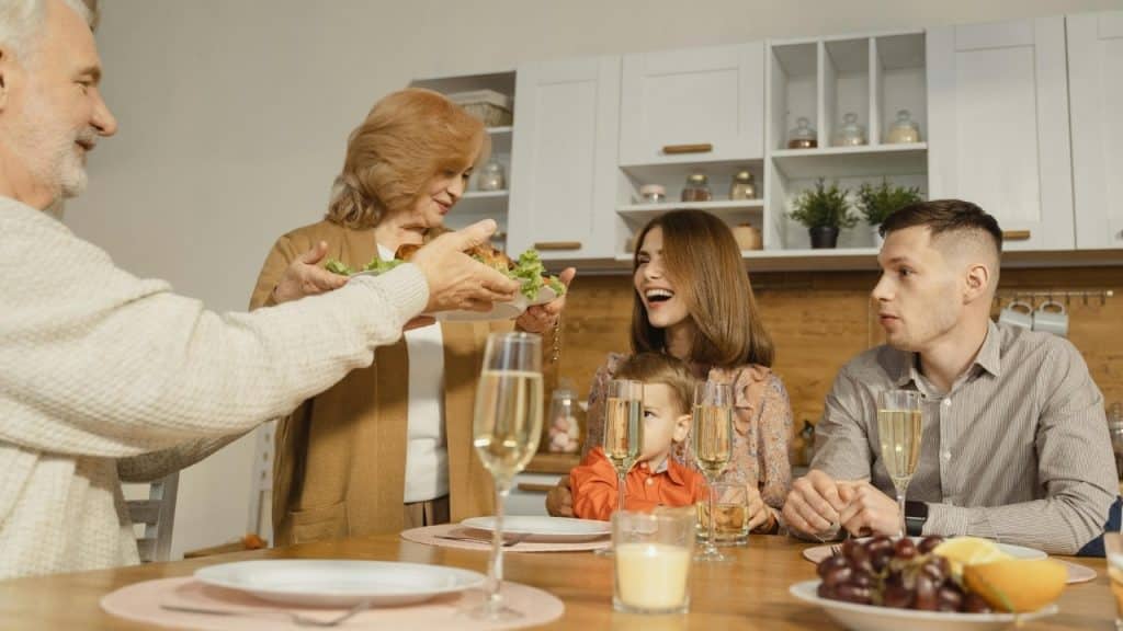 A family group gathered around a wooden dining table while a woman serves a platter.