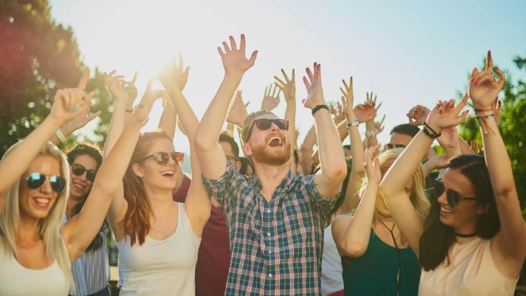 A crowd of people with their hands raised cheer outdoors under a bright sun.