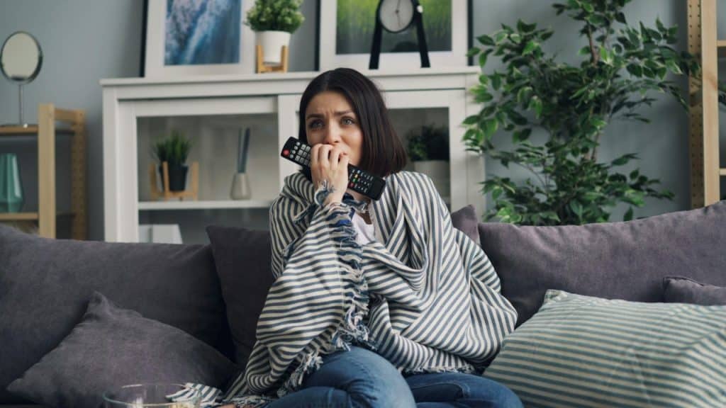 A woman wrapped in a blanket sits on a sofa holding a remote and biting her hand.