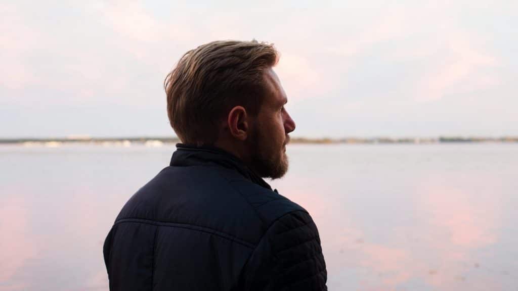 A bearded man in a dark jacket looks out over a calm body of water.