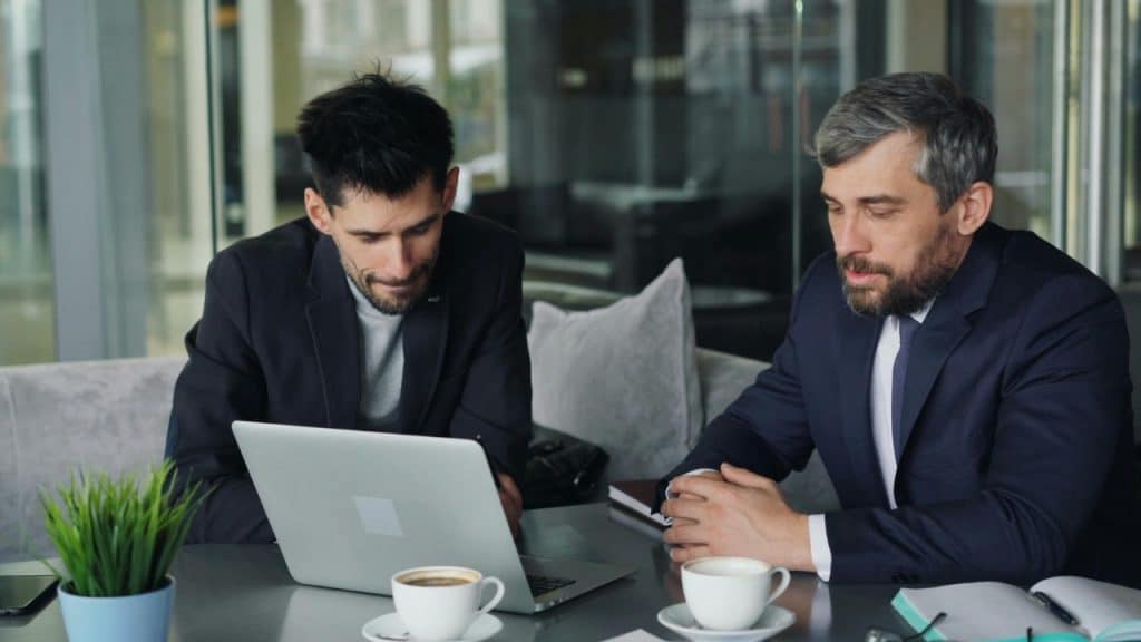 Two men in suits sit at a table looking at a laptop with coffee cups.