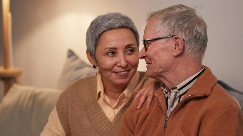 An older man and woman sit close together on a sofa, looking at each other.