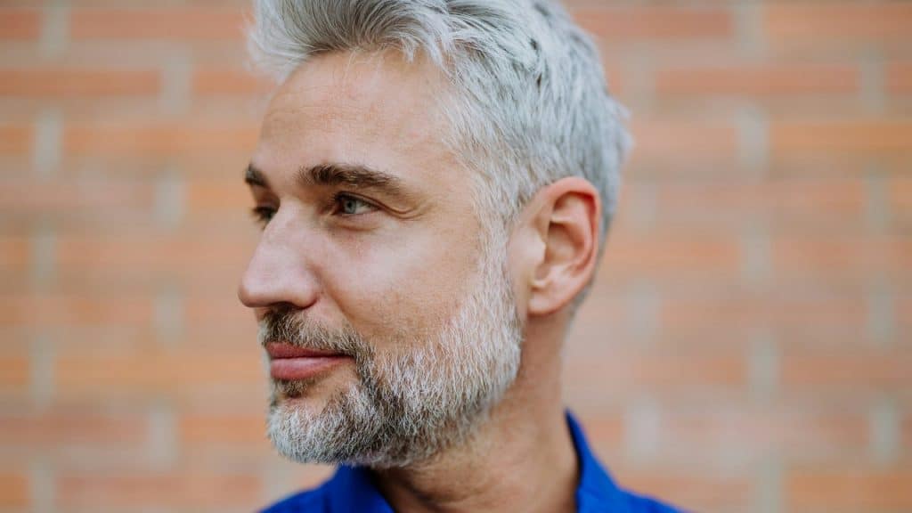 A close-up profile of a man with grey hair and a beard against brick.
