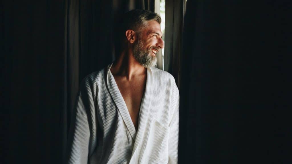 A man with a grey beard wearing a white robe stands smiling by a window.