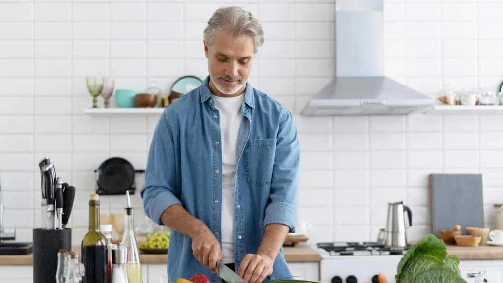 A man in a denim shirt stands in a kitchen cutting vegetables on a board.