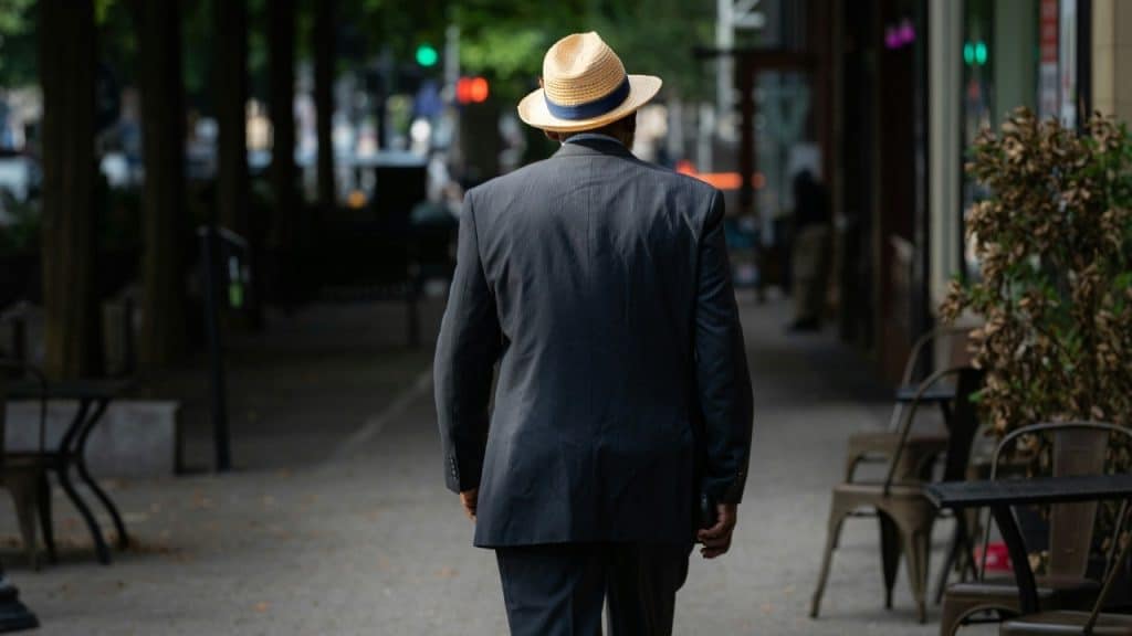 A man in a grey suit and straw hat walks down a tree-lined city sidewalk.
