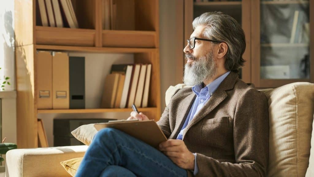 A bearded man with glasses holds a clipboard and looks thoughtfully out a window.