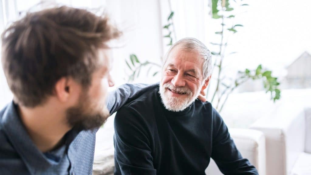 A smiling older man with a grey beard looks at a younger man beside him.