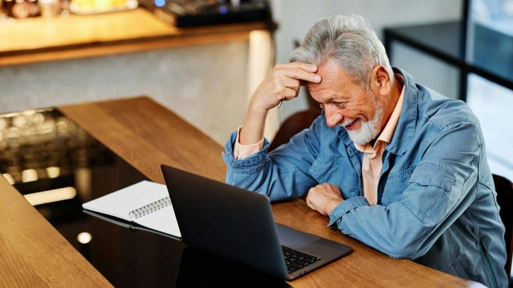 A grey-haired man smiles while looking at a laptop at a wooden table.