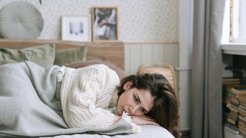 A woman lies on a bed wrapped in a blanket, looking sad
