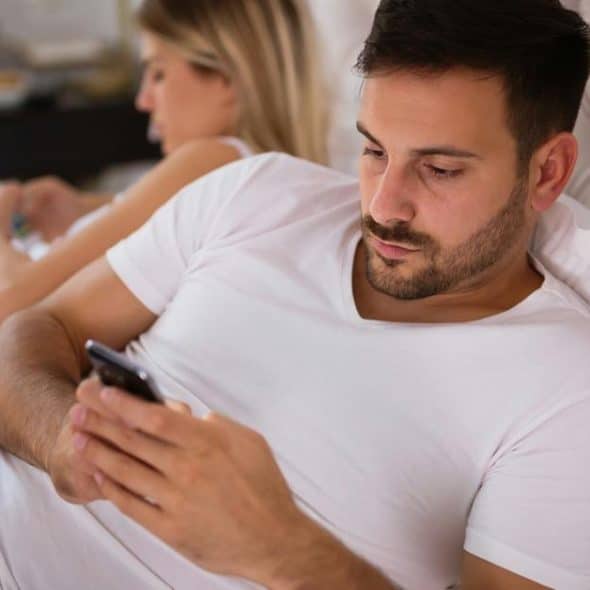 A couple lies on a bed while both look at their phones