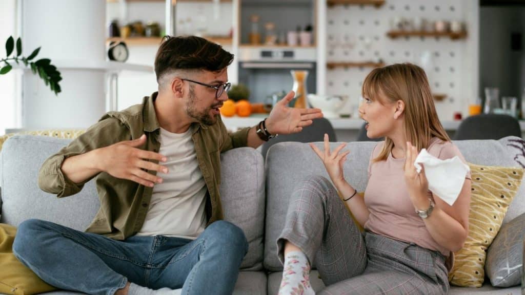 A man and woman sit on a grey sofa and gesture during a conversation.