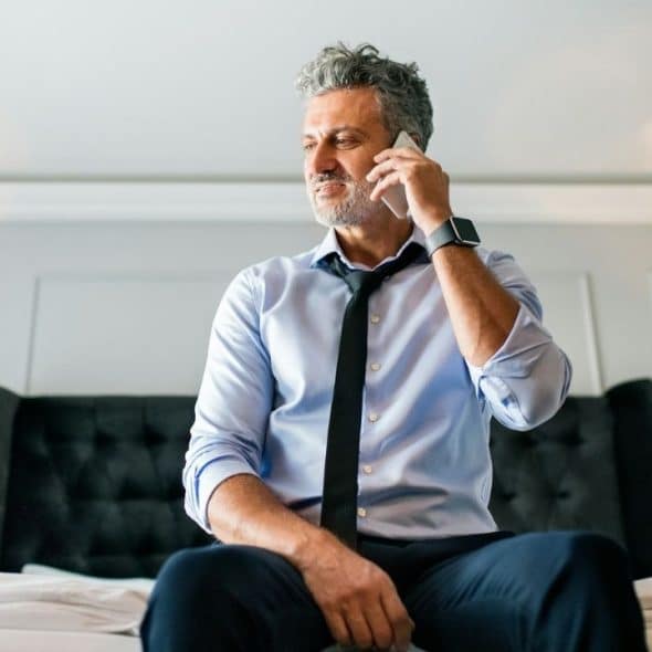 A man with grey hair sits on a bed while talking on a mobile phone.