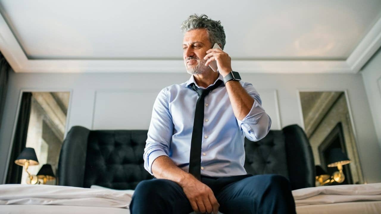 A man with grey hair sits on a bed while talking on a mobile phone.