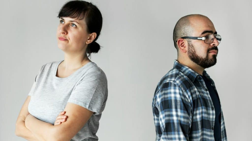 A woman and a man stand back-to-back against a plain grey background.