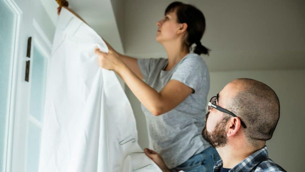 A woman hangs white curtains while a man in glasses watches from a lower angle.
