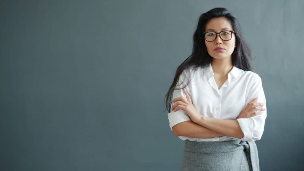 A woman with long dark hair and glasses stands with her arms crossed.