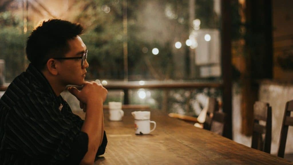 A man with glasses sits alone at a long wooden table in a restaurant.