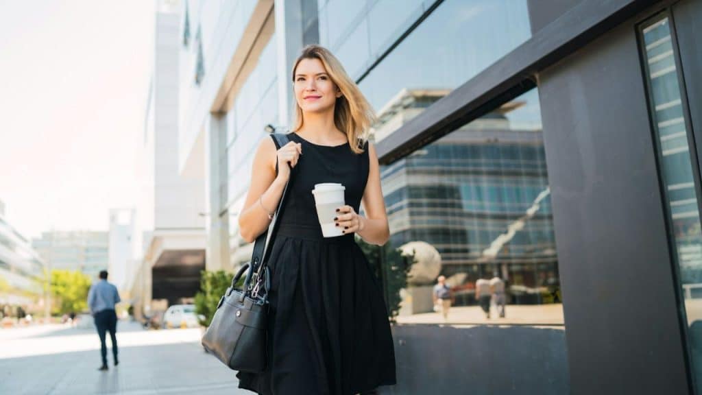 A woman in a black dress walks outside while holding a white coffee cup.
