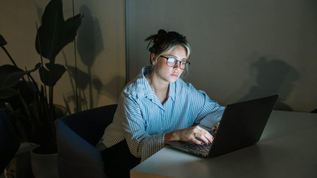 A woman wearing glasses and a striped shirt works on a laptop at night.