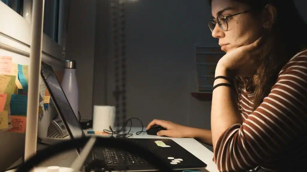 A woman wearing glasses working on a laptop at a desk with sticky notes on the wall.