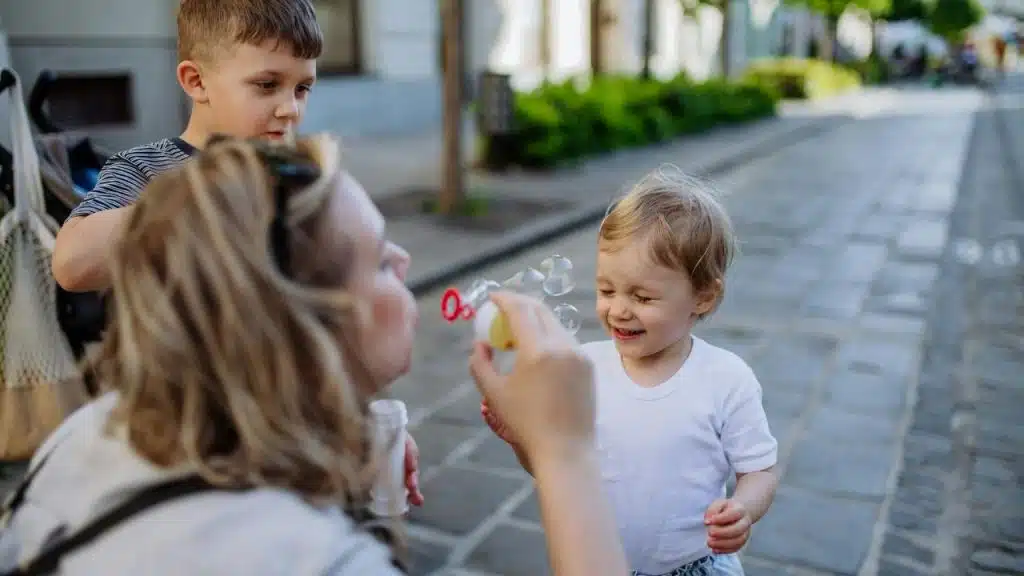 A woman blowing bubbles for two young children on a cobblestone street.