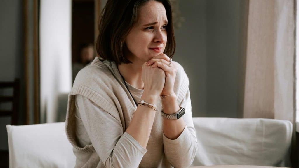 A worried woman sits indoors with her hands clasped near her face.