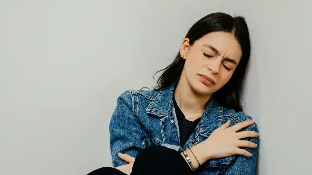 A distressed woman in a denim jacket sits against a wall with her eyes closed.