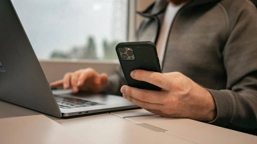 A person using a smartphone while working on a laptop at a desk.
