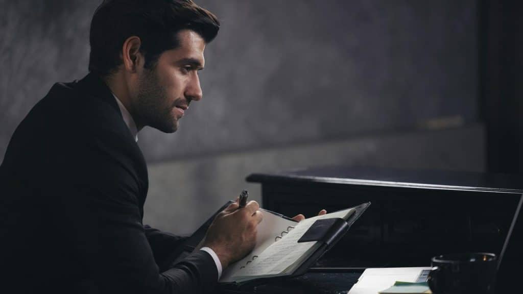 A man in a suit writing in a notebook beside a piano.
