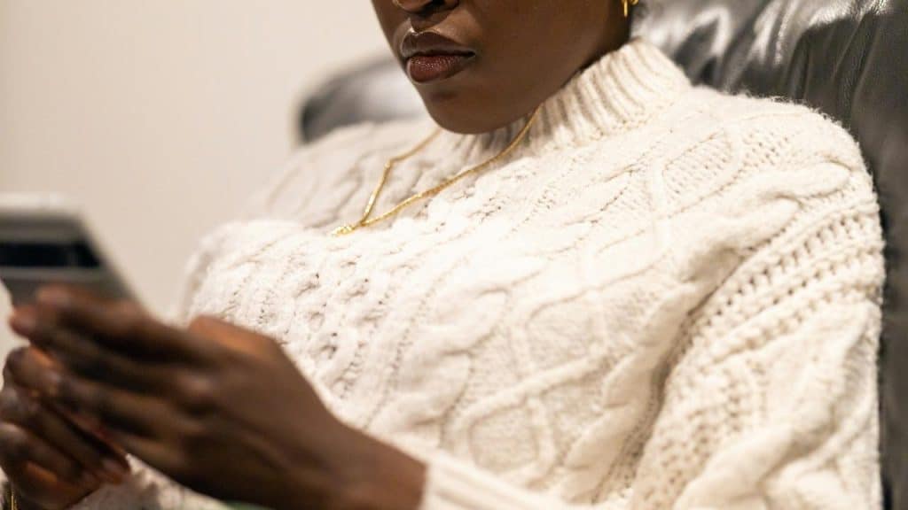 A person in a white knit sweater looking at a smartphone while seated.
