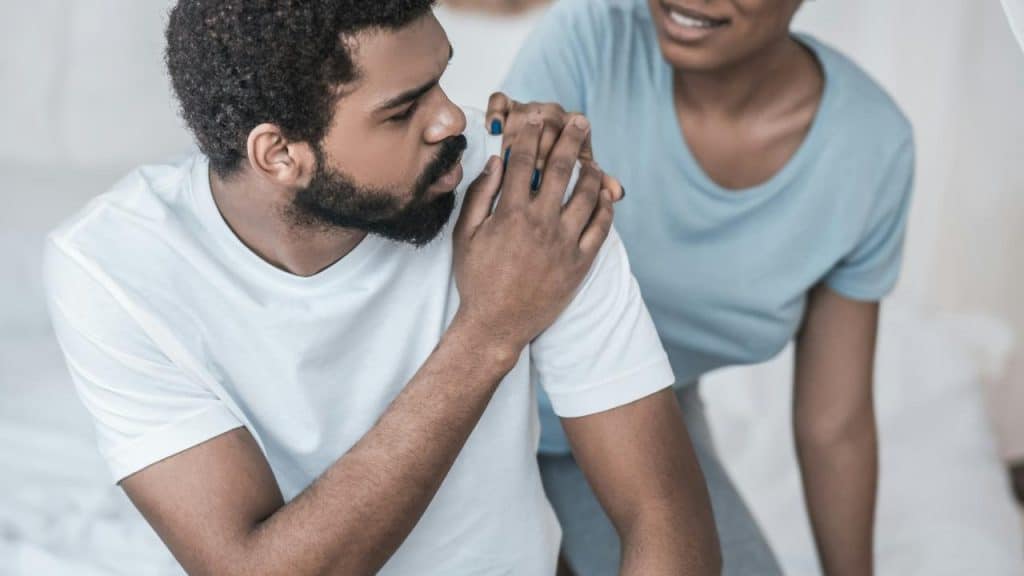 A worried man sits while a woman comforts him with a hand on his shoulder.