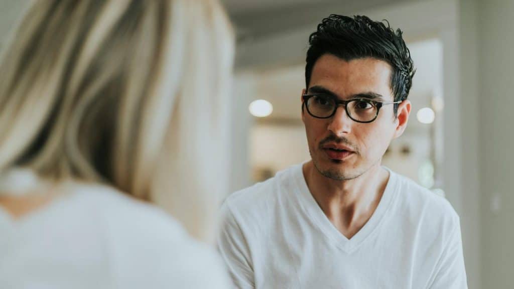 A man wearing glasses and a white shirt talks with a woman in an indoor setting.