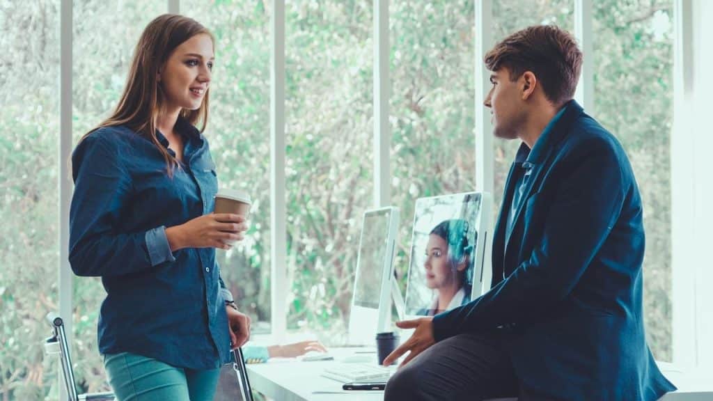 A woman holding a coffee talks with a man sitting on a desk in an office.