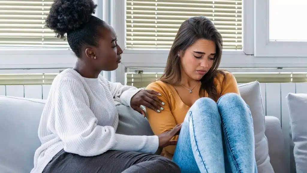 A woman comforts a sad friend sitting on a couch.