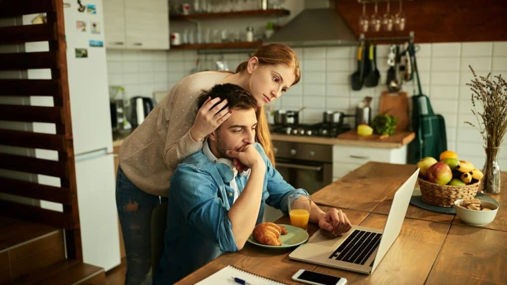 A woman comforting a man as he works on a laptop at a kitchen table.