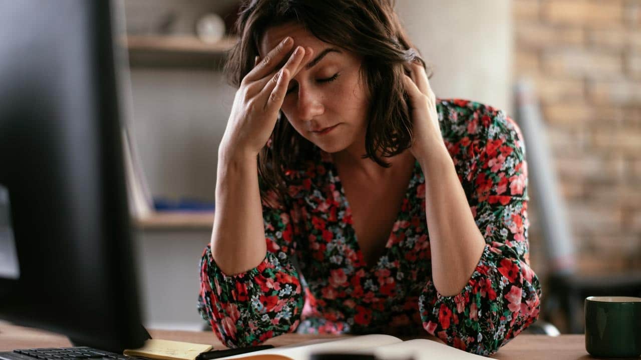 A woman sits at a desk with her hand on her forehead