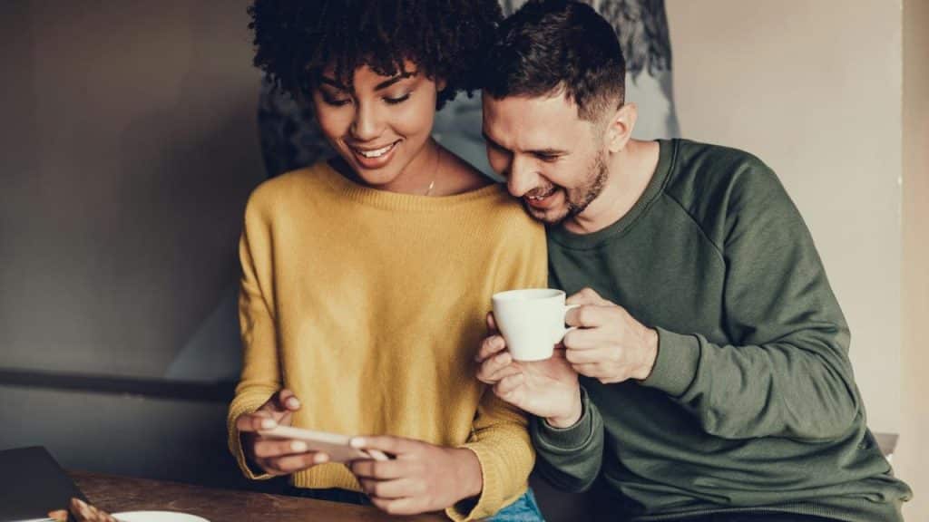 A couple smiling together while looking at a phone and holding a coffee cup.