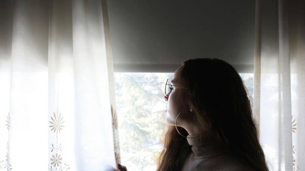 A woman looking out a window through sheer curtains.