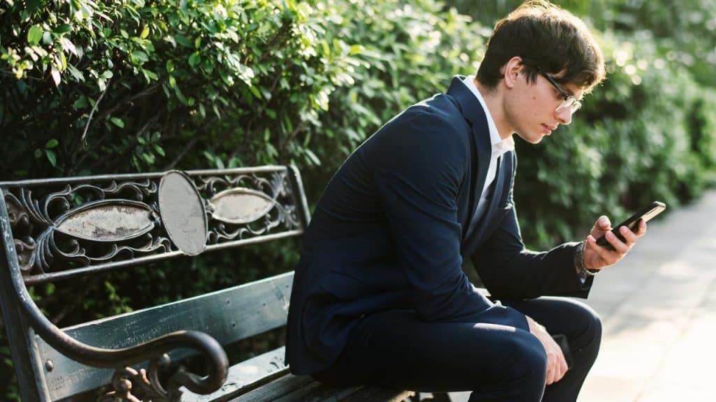 A man in a suit sitting on a park bench and looking at his phone.