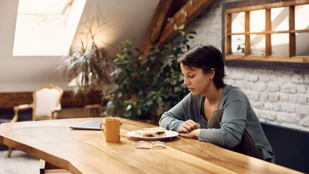 A woman sitting at a wooden table, looking down at a plate and mug.