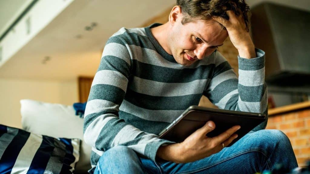 A man sitting indoors and looking at a tablet with his hand on his head.