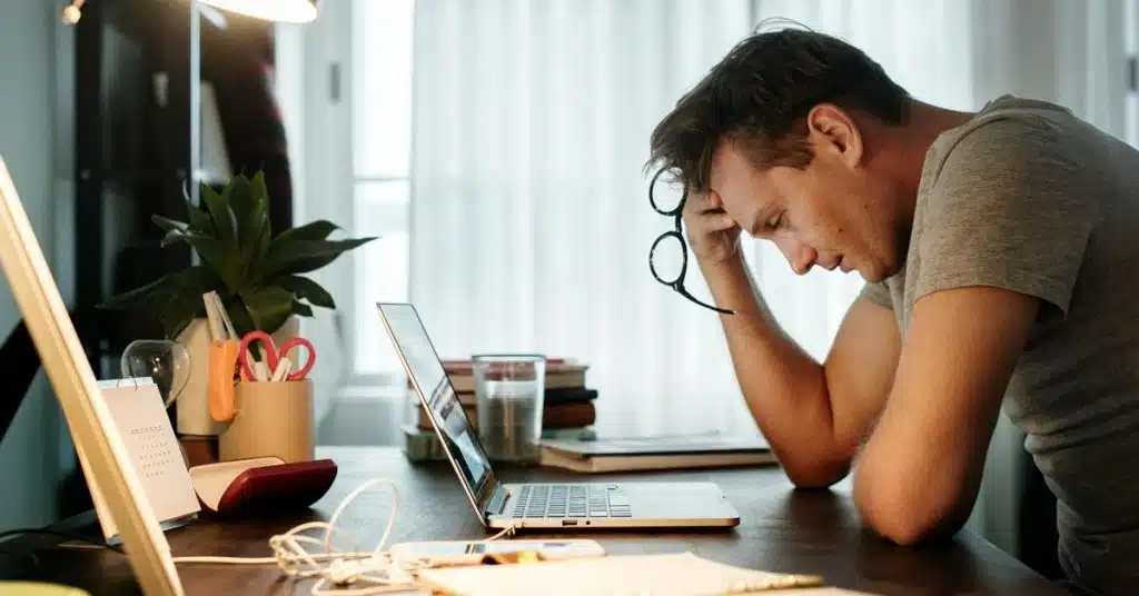 A person sits at a desk holding glasses while looking at a laptop.