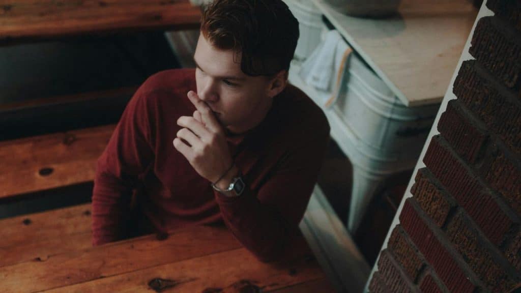 A man sitting at a wooden table, looking away thoughtfully with his hand near his mouth.