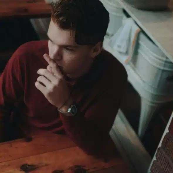 A man sitting at a wooden table, looking away thoughtfully with his hand near his mouth.