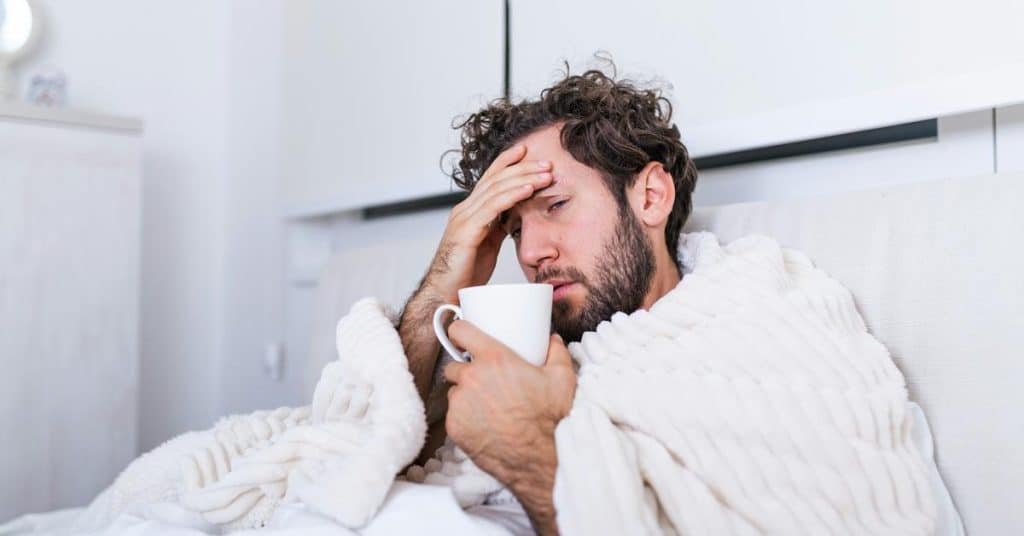 A man wrapped in a blanket holds a mug and rests his hand on his forehead.